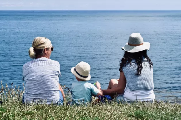 Två kvinnor och ett barn sitter på en gräsmatta och tittar ut över havet. Foto.