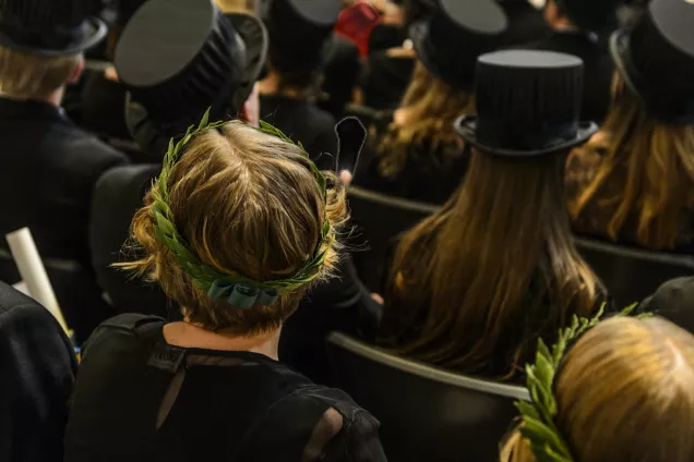 Personer sitter på stolar under en doktorspromotionen, med lagerkrans eller hatt på huvudet. Foto.